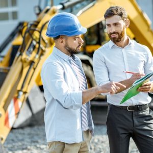 A man in a blue hard hat talks to a man in professional attire with paper and a pencil on a construction site.