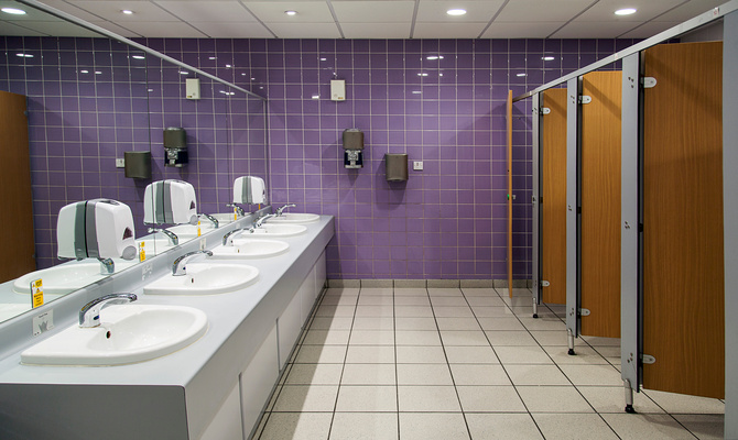 A public restroom features a long mirror and purple tile on the far wall. The stalls have wooden doors.
