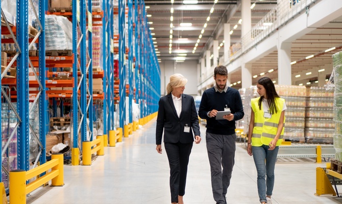 A man walks down the aisle of a warehouse, and a woman walks on either side of him. He is holding a tablet.