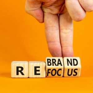 A person uses two of their fingers to hold wooden blocks in a position to simultaneously spell "rebrand" and "refocus."