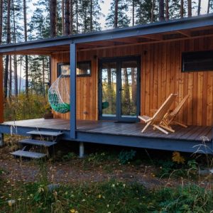 A tiny home made out of stained wood in the middle of a forest. A hanging chair and two wood chairs are on the porch.