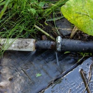 A close-up of a water pipe with two different pipes connecting and water spraying out from leaks at the connection.