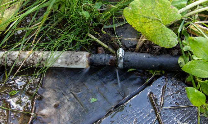 A close-up of a water pipe with two different pipes connecting and water spraying out from leaks at the connection.