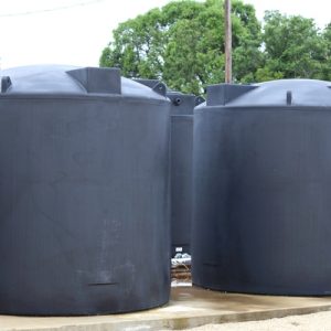Four large black municipal water tanks with sealed tops are resting on a concrete foundation outdoors.