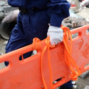 Two people wearing blue pants and white gloves carry an orange rescue stretcher through an area with a fallen building.
