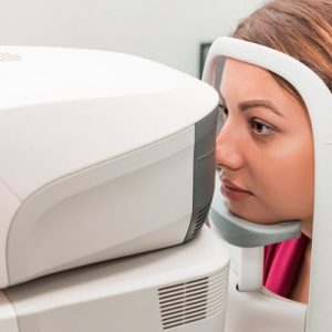 A brown-haired woman in a red shirt using a chin rest while looking into a large white eye exam machine.