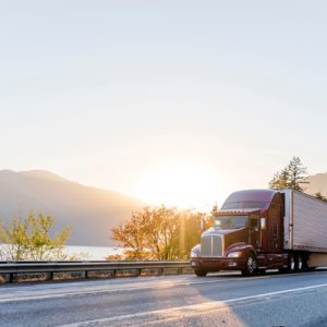 A burgundy semi-truck transporting commercial cargo is driving on the road along the river with sunshine in the background.
