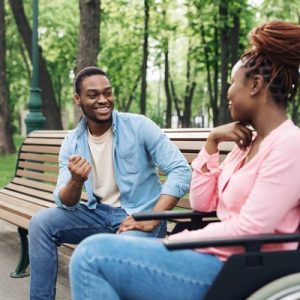 A young couple sitting in a vibrant park with tall trees. One person is on a bench and the other is in a wheelchair.