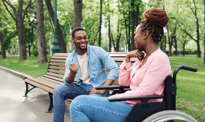 A young couple sitting in a vibrant park with tall trees. One person is on a bench and the other is in a wheelchair.