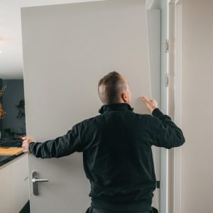 A person wearing a black jacket installs a new white door with a silver handle into a door frame in a kitchen area.