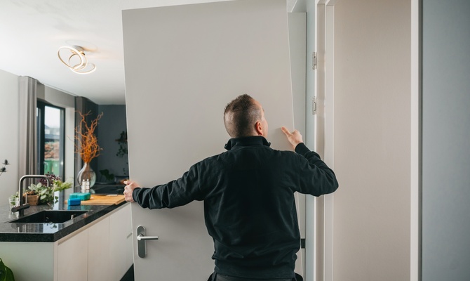 A person wearing a black jacket installs a new white door with a silver handle into a door frame in a kitchen area.