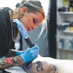 A tattoo artist with a wrapped cordless rotary machine focusing as they tattoo a design on a client's shin.