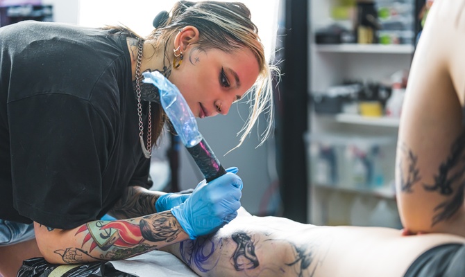 A tattoo artist with a wrapped cordless rotary machine focusing as they tattoo a design on a client's shin.