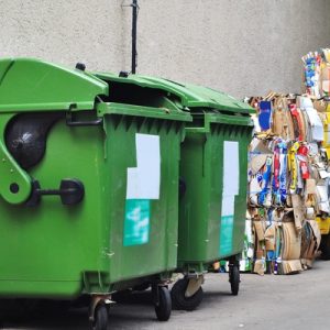 An alley behind a business with two green trash hoppers and stacks of compacted cardboard to the right of the hoppers.
