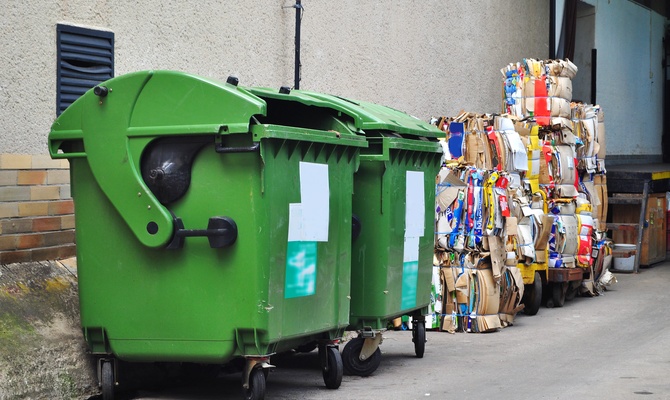 An alley behind a business with two green trash hoppers and stacks of compacted cardboard to the right of the hoppers.