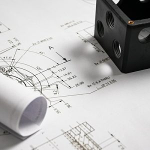 A black plastic box with threaded holes on top of a paper featuring technical drawings and designs.