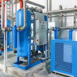 An air compressor system comprised of blue, gray, and red machinery and pipes in a warehouse with cement floors.