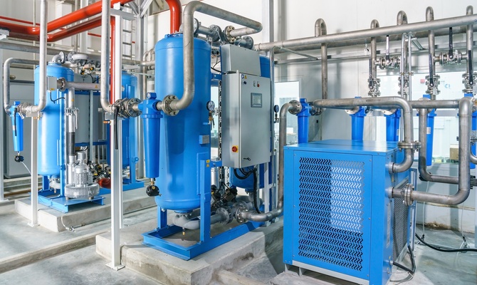 An air compressor system comprised of blue, gray, and red machinery and pipes in a warehouse with cement floors.