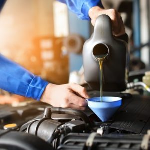 A mechanic in a blue jumpsuit pours automotive oil into the engine of a car through a light blue funnel.