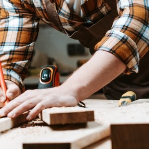 A woodworker uses tools to measure a wooden plank in their woodworking workshop. There are other tools next to them.