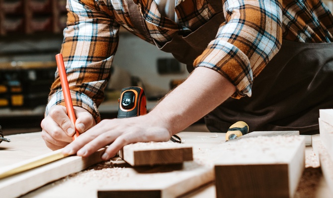A woodworker uses tools to measure a wooden plank in their woodworking workshop. There are other tools next to them.