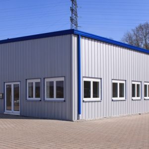 The exterior of a gray steel building with blue accents, several windows, a garage door, and a brick driveway.