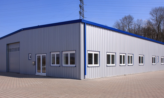 The exterior of a gray steel building with blue accents, several windows, a garage door, and a brick driveway.