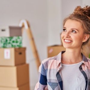 A woman is smiling in her new home. There are boxes of her belongings and indoor plants in the background.
