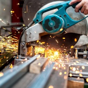 An industrial worker uses a metal circular saw to cut through a thick sheet of metal in a metal workshop.
