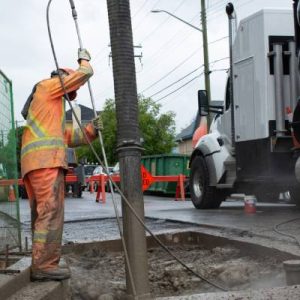 A worker stands over an excavation hole and plunges a long tube for a water jet next to a suction tube for hydro excavation.