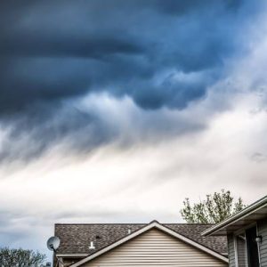 Some homes sit underneath a sky that is full of dark thunder clouds. Past the homes, there are trees in the distance.