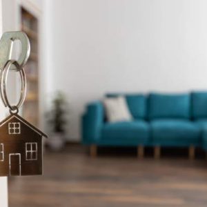 An out of focus apartment living room shown through an open door, where a key with a house keychain is in the lock.