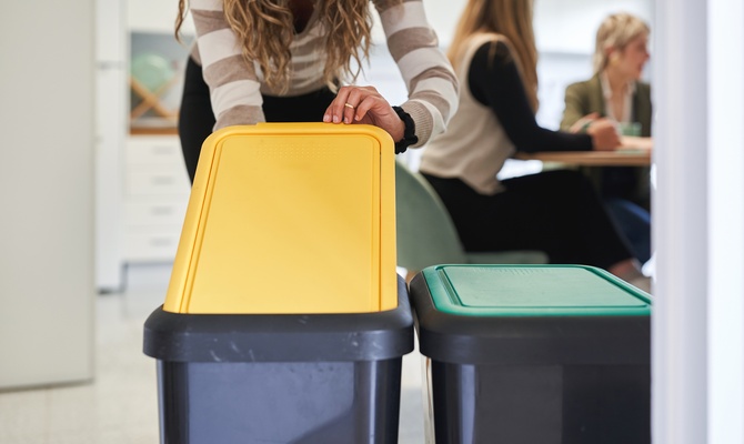 A close-up shows a person wearing a striped sweater placing something into a black garbage can with a yellow lid.