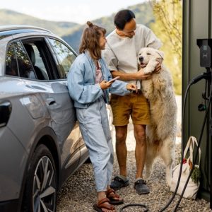 A young man and woman stand next to a large dog as they charge a silver electric vehicle on a sunny day.