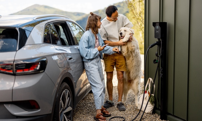 A young man and woman stand next to a large dog as they charge a silver electric vehicle on a sunny day.