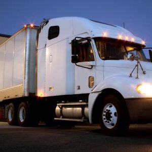 A white semi truck with a white trailer is backed into a warehouse docking station at dusk. The truck's lights are on.