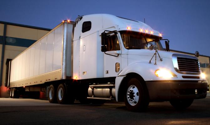 A white semi truck with a white trailer is backed into a warehouse docking station at dusk. The truck's lights are on.