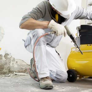 A man in a white hard hat, mask, and gloves adjusting a yellow air compressor on a construction site.