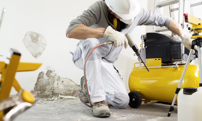 A man in a white hard hat, mask, and gloves adjusting a yellow air compressor on a construction site.