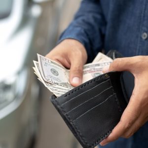 A person wearing a denim shirt holds open a black wallet, revealing multiple bills of money while standing next to a car.