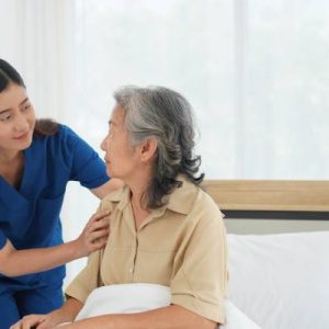 A young woman caregiver is taking care of an older Asian woman patient. The patient is lying in the bed.