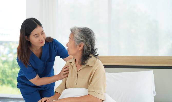 A young woman caregiver is taking care of an older Asian woman patient. The patient is lying in the bed.
