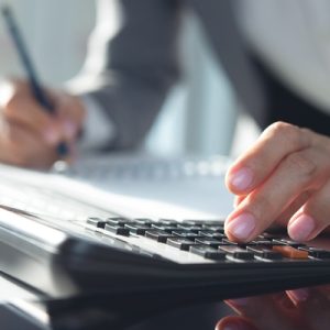 A close-up of a person pressing a button on a calculator with one hand while writing on a notepad with their other hand.