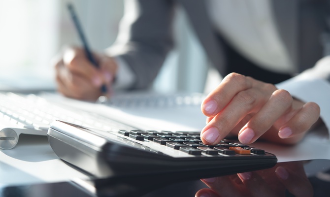 A close-up of a person pressing a button on a calculator with one hand while writing on a notepad with their other hand.