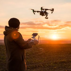 A person in a black knit hat and a brown jacket flies a drone above agricultural fields at sunset.