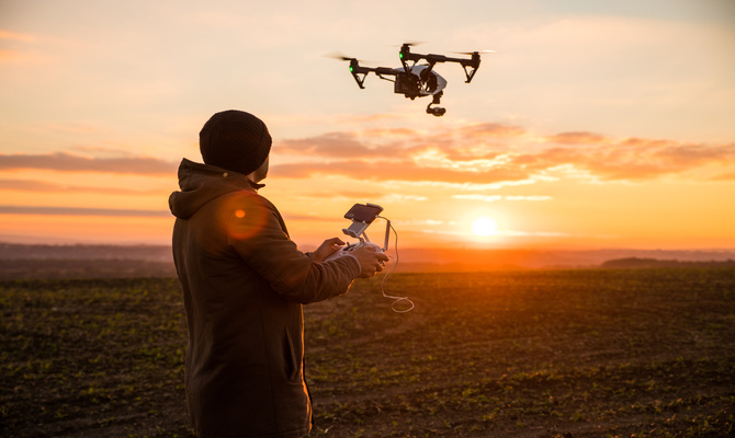 A person in a black knit hat and a brown jacket flies a drone above agricultural fields at sunset.
