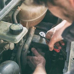 A man, dirty from grease and oil, leans over the hood of his car, using a wrench to tighten a bolt.
