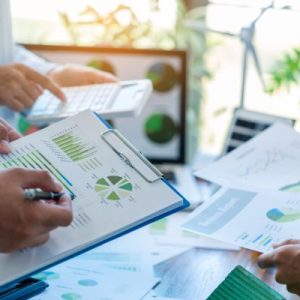 A round table with multiple people standing and looking at different pages of charts and graphs, and a calculator.