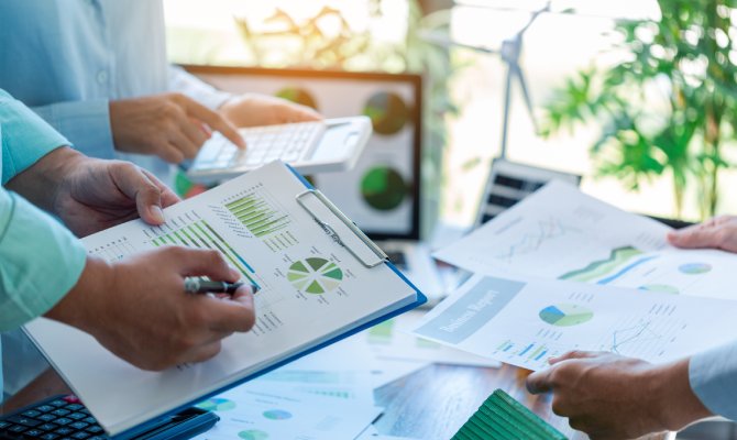 A round table with multiple people standing and looking at different pages of charts and graphs, and a calculator.