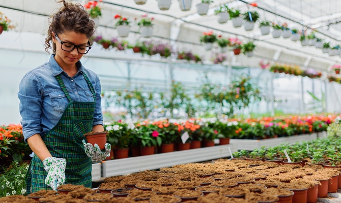 A woman in a large building is wearing an apron and gloves and works on some small pots containing different plants.